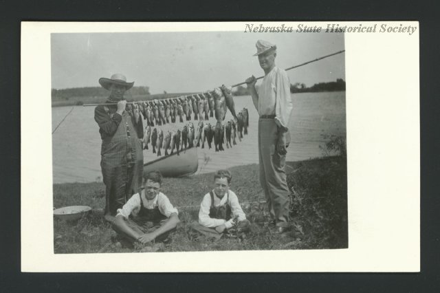 Two men with two boys hold up their catch of fish. At least forty fish are strung on a cane pole and stringer for this trophy shot. A lake and row boat are behind the people. The same fish are in 141-8.