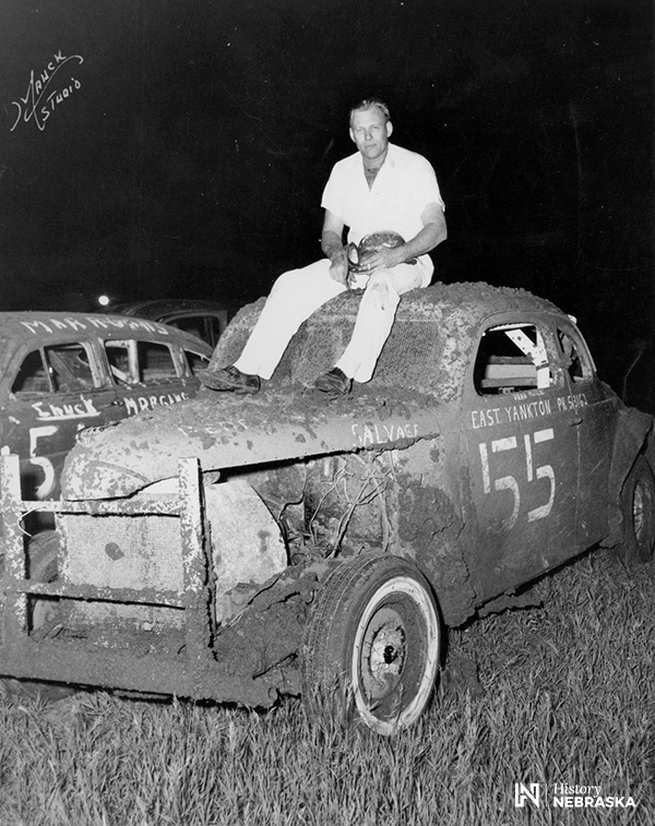 man sitting atop mud-spattered 1930s-model stock car, circa 1950s
