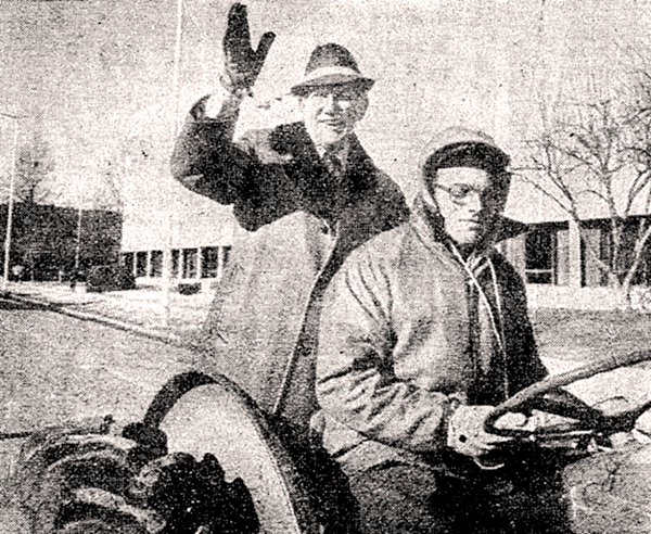 Gov. Jim Exon waving from the back of a tractor driven by another man.