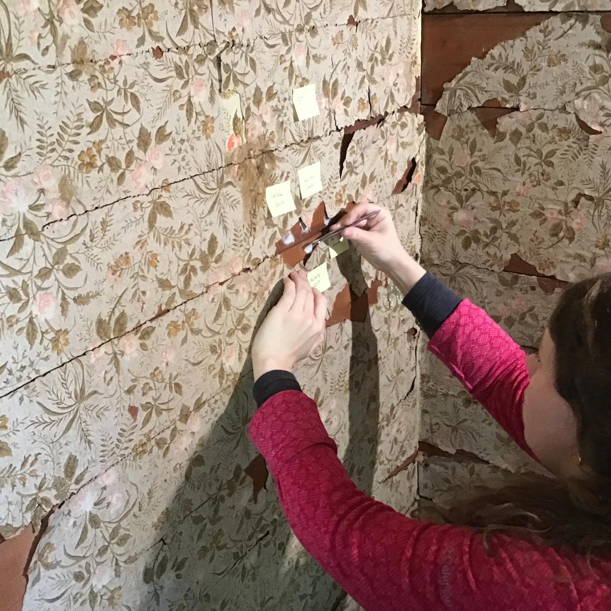 Hilary LeFevere is sitting on the floor, adhering small pieces of Japanese tissue to the original wall boards, using sticky notes to label different materials.