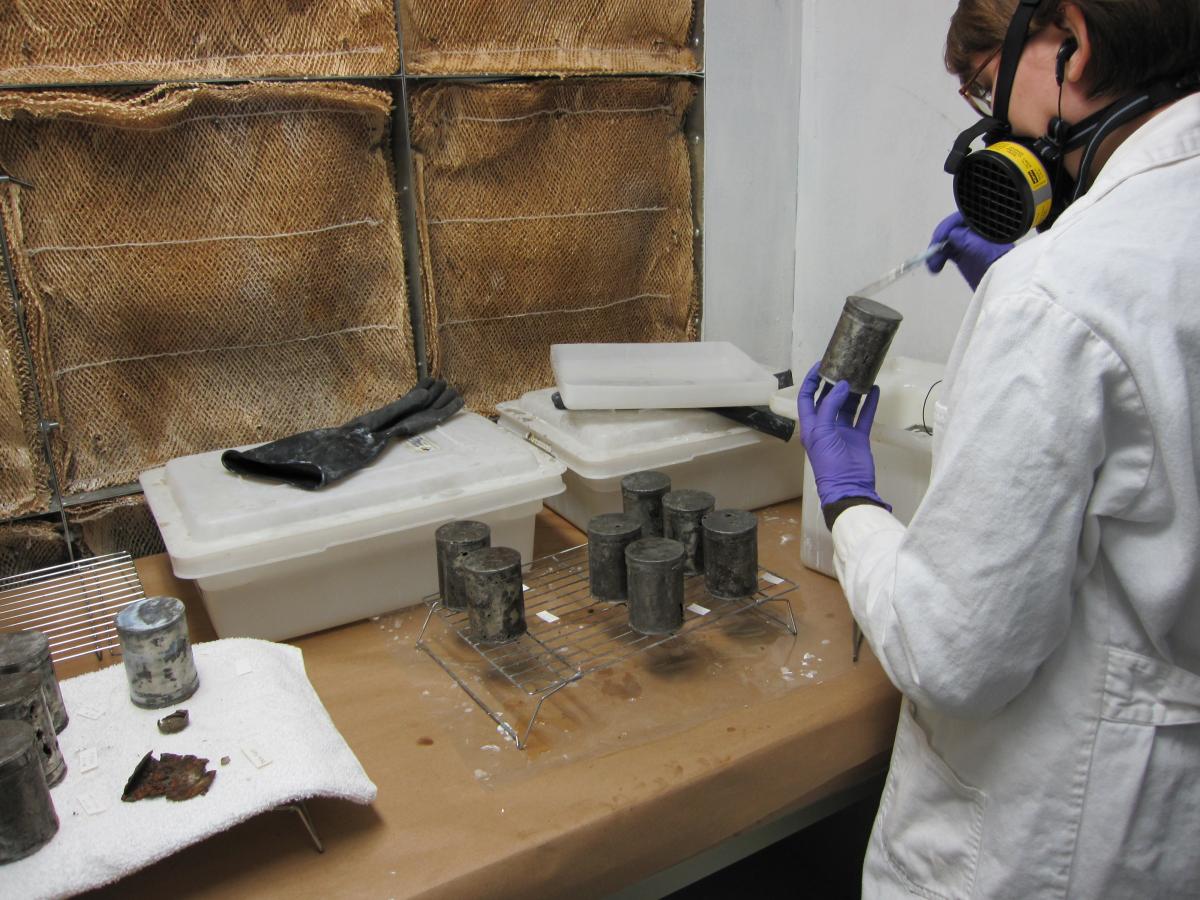 woman in white lab coat and respirator holds can in left hand and applies coating with a brush in the right hand.  Tanks of solvent and additional cans are in the image.