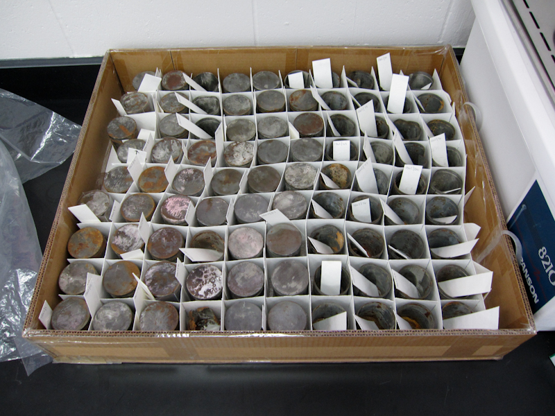 Box with 90 yeast cans, each in separate divider with paper label. Metal showing corrosion and losses