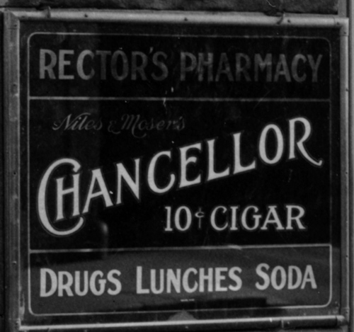 Detail of the corner drug store. A sign that reads: Rector's Pharmacy, Chancellor 10 cent cigar, Drugs, Lunches, Soda