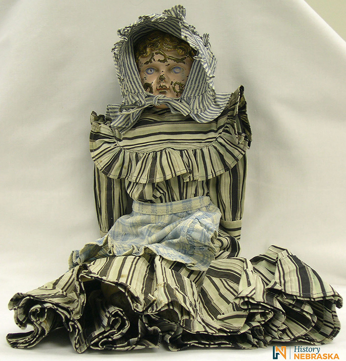 Doll wearing a brown and white striped dress, blue and white apron, and blue and white bonnet