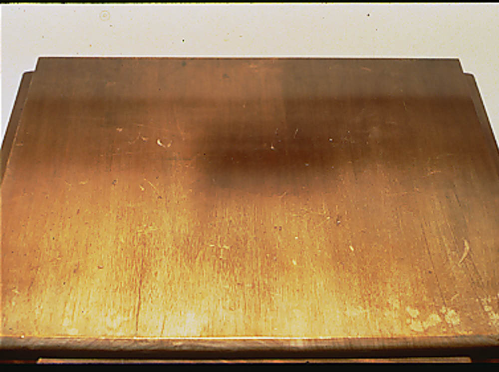 Photograph of a table top. The front half of the table has been exposed to light and has bleached and the surface coating has cracked and pealed. There are darker rectangles in the upper right corner where a lamp or other object blocked the light.