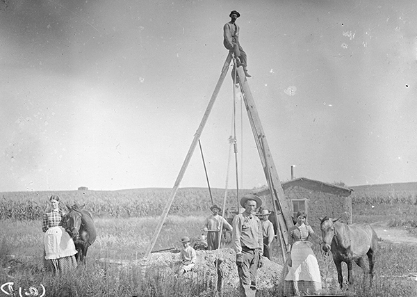 Man perched on a large wooden tripod over well and pile of dirt. Other people standing around; sod house and cornfield in background.