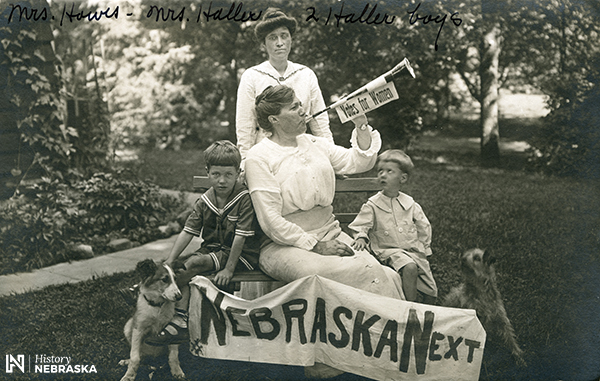 Woman blowing a horn labeled "Votes for Women." Banner reads, "NEBRASKA NEXT"