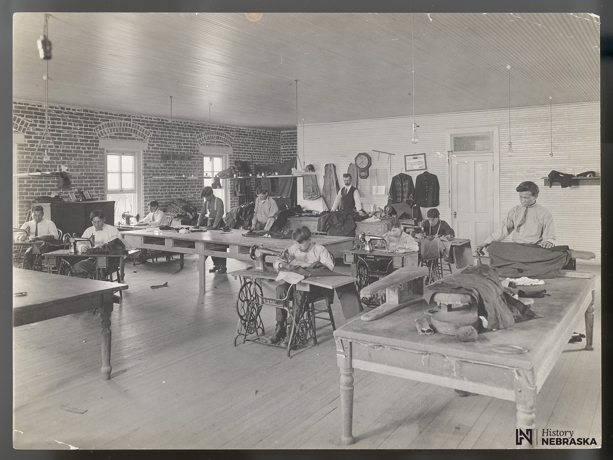 Young men working at sewing machines inside a large room at the Genoa Indian School