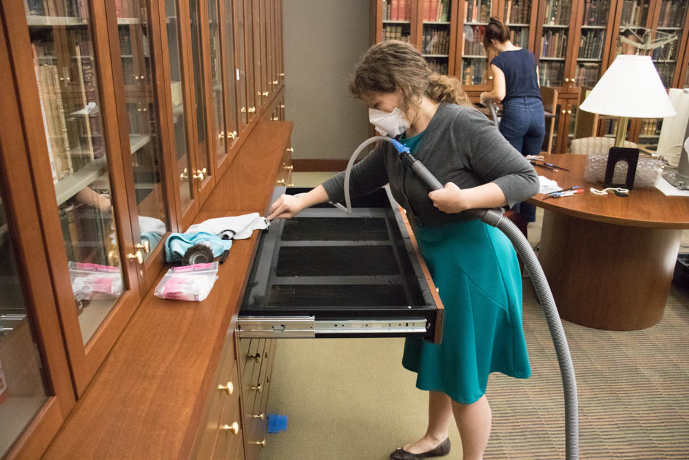 Hilary LeFevere, Paper Conservator, cleans out one of the drawers after the books have been removed.  Right: Vonnda Shaw, Conservation Technician, vacuums the spines of a shelf worth of books.