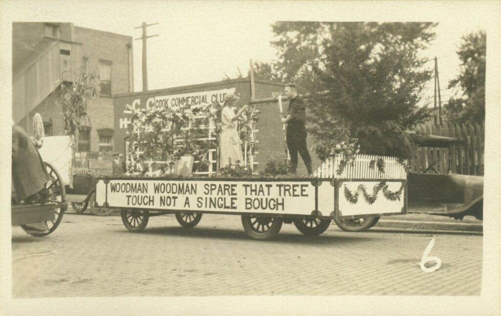 A parade float with "Woodman Woodman Spare That Tree Touch Not a Single Bough" written on the side. A man holding an axe and a woman in pioneer dress stand on the float.