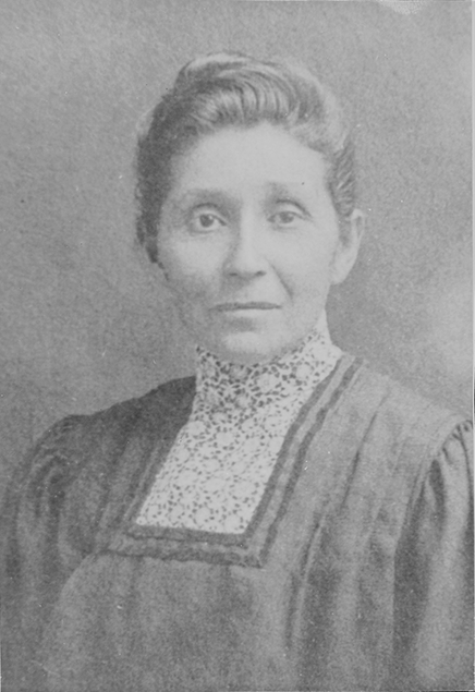 Black and white portrait of a woman with her hair pinned up, wearing a high-collared floral blouse and a dark dress, looking directly at the camera.