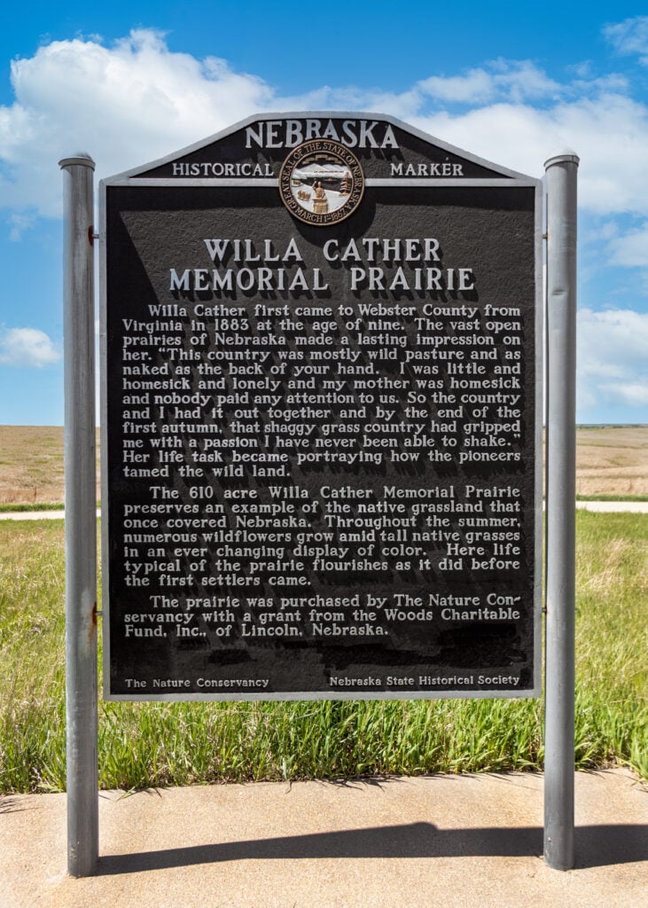 Nebraska historical marker for Willa Cather Memorial Prairie, detailing Cather’s connection to the land and the prairie’s ecological restoration, with blue sky and grassland in the background.