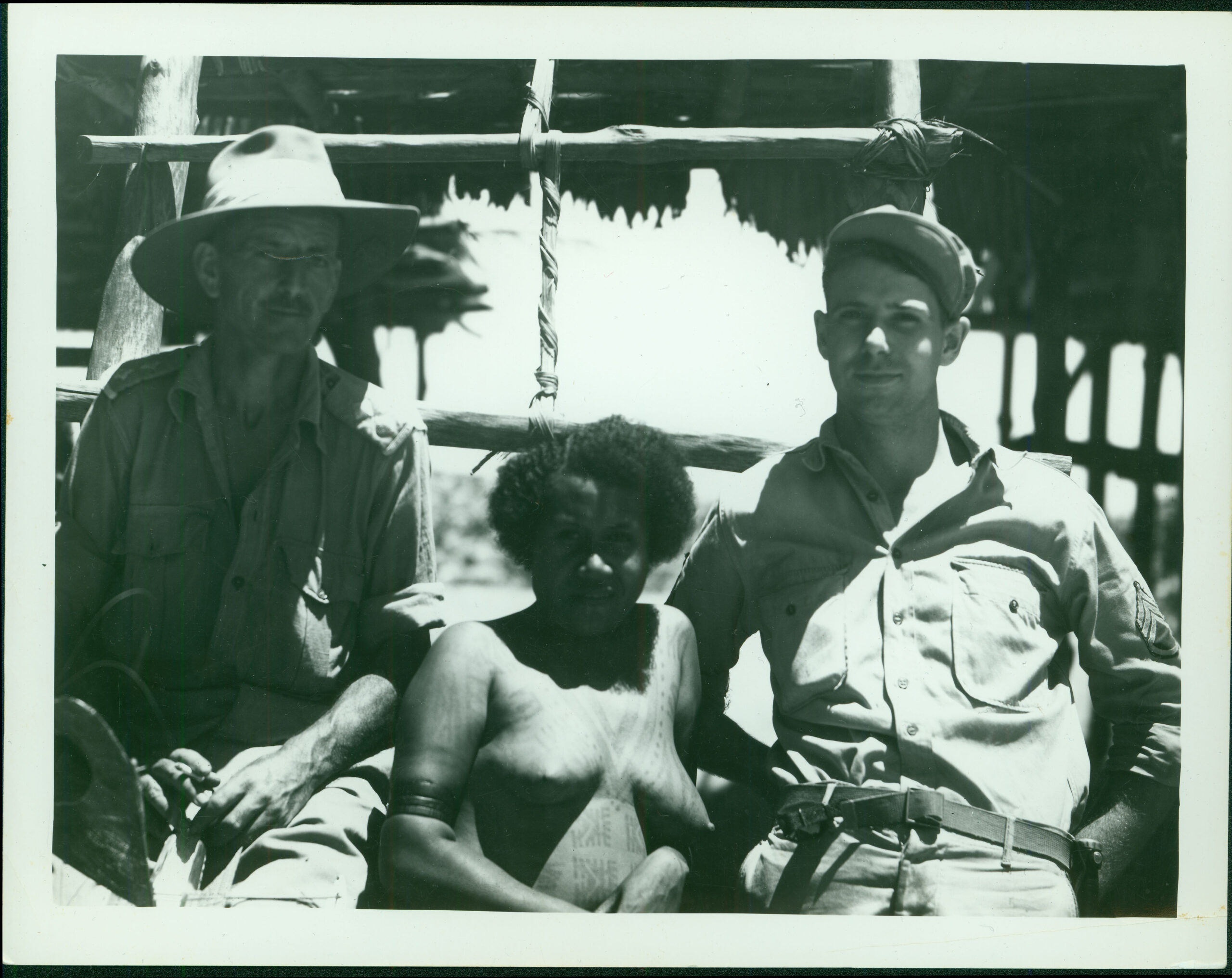 While visiting a native village Robert Merchant and an Australian Army Lieutenant pose in front of a village hut with a native woman. A note from Merchant on the back of the photo reads , “Dad, was it hot this day”.