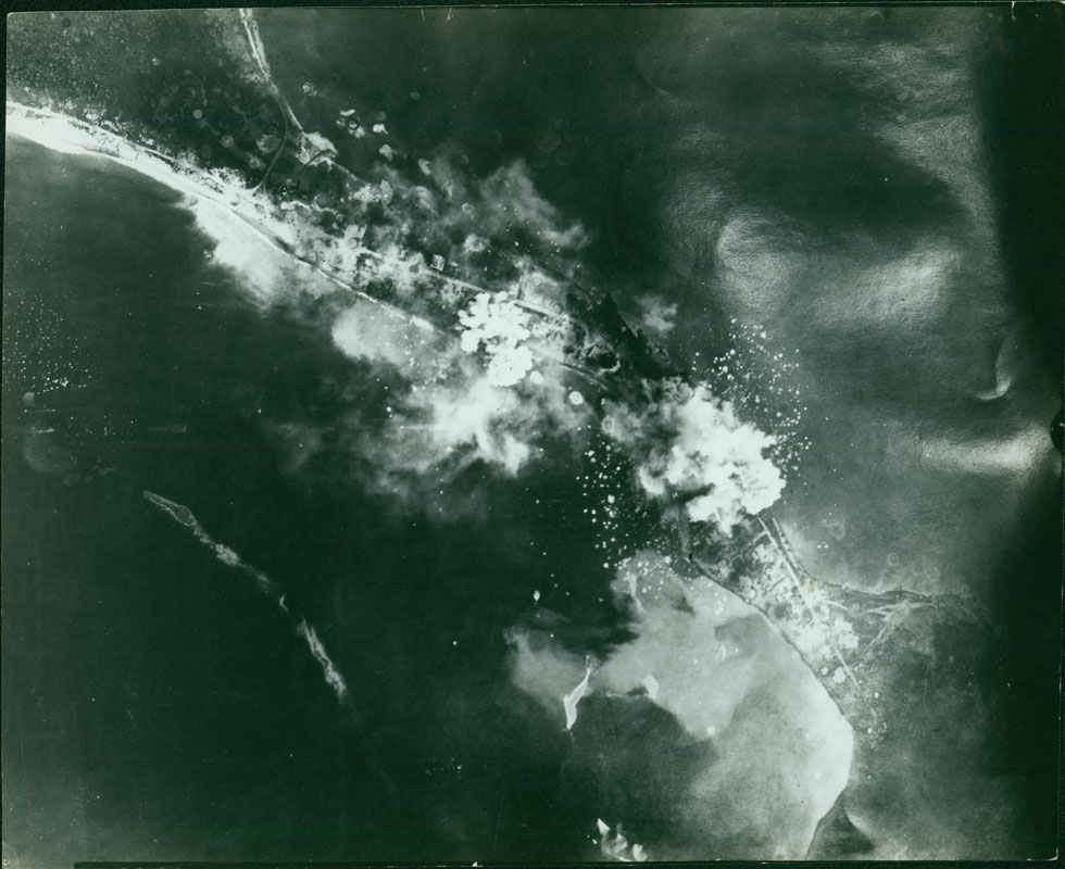 This shows an aerial view of bombs exploding over land and water. The back of the photo reads "Salamaua Peninsula" Salamau was originally intended by the The Japanese originally intended Salamaua as a staging post for an assault on Port Moresby, because taking control of the Australian territory of New Guinea was a major aspect of Operations Mo, Japan’s overarching plan to isolate Australia and New Zealand from the United states. However, the plan to take Port Moresby was abandoned after several failed attacks. Instead, the Japanese converted the Salamaua port into a major supply base.