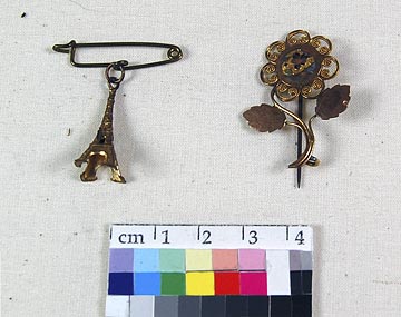 Brooch (NSHS 7144-121 ) Brooch consisting of two pieces tied together. The upper pin consists of a gold colored rosette with a center floral motif. It has a straight pin clasp soldered on the back. The Tower piece is a small gold-colored Eiffel Tower with a safety pin attached to the top of the tower. 