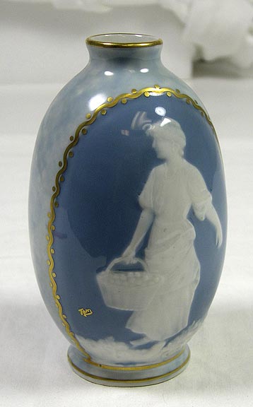 Limoges vase with a figure of a woman with a basket in one hand and the opposite arm extended. Gold trim surrounds that opening and frames the cameo of the woman. On the base it says: "MT Limoges". Below the cameo signed in gold is the name "Tom." (NSHS 7144-124)