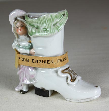 Ceramic vase that consists of a boot with a painted buckle and a figurine of a girl in period costume that serves as a handle. Attached tag says: "FROM ENGHIEN, France." (NSHS 7144-168)