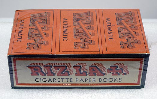 Cigarette papers (NSHS 7144-179) Cellophane-wrapped carton of Riz-La+ cigarette papers has twenty-one books of papers. The books are orange and yellow and the packet has a green rim.  The side of the carton says: "Riz La +, Cigarette Paper Books"  The top of the carton says: "Automatic Gummed, The Only Genuine LA + Wheat Straw Cigarette Paper".  The bottom of the packet says: "Thanks for the friendship trains. Back to you again since 1940, French Made Riz La+, Cigarette Paper Books, Presented by L. LACROIX Fils, ANGOULEME, Paper Makers Established 1728, Ready to resume shipments to U.S.A."