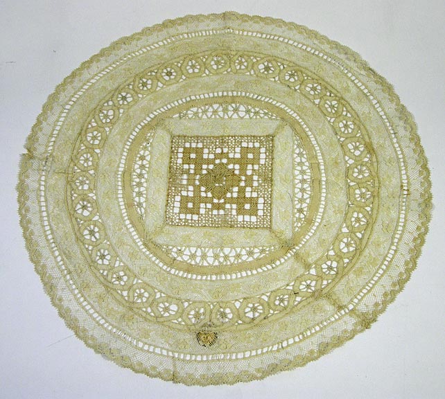 Doily (NSHS 7144-205) Lace doily with a geometric design in the center square that is surrounded by lace with floral patterns.