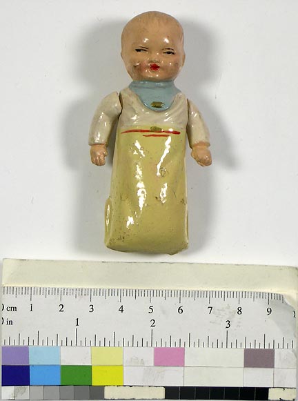 Doll (NSHS 7144-25) Composition baby doll in a sleeper with a red painted belt, cream top, and light blue bib. Arms are movable and held to the trunk with a string. Inscriptions: Letters "NIL" cast in raised letters on back of neck. Doll is 3.25" (8.2 cm) long. 