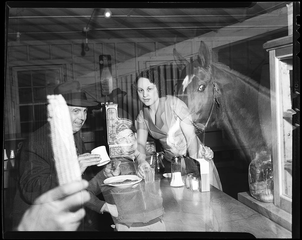 In this image from 1939, Dewell mistakenly captured two shots on the same negative. One shot shows a man holding the reins of a horse, while the other image features a woman serving food to a customer. The combination results in a humorous image in which it appears that a horse is visiting for a bite to eat. (RG3882.PH000002-000748)