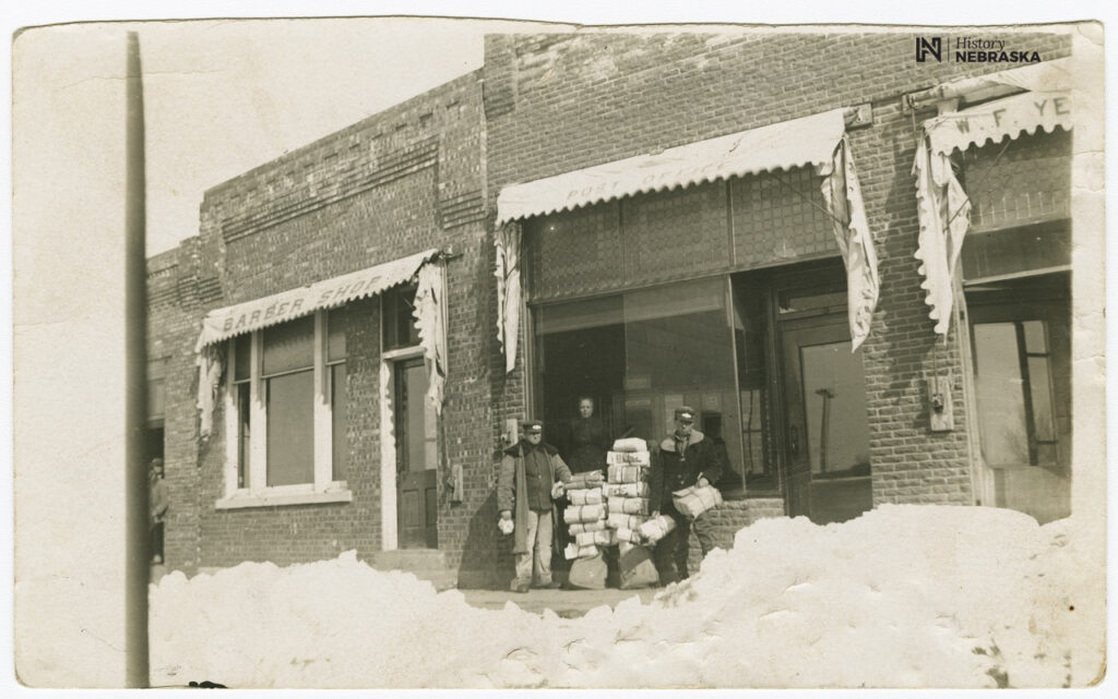 Black and white photograph of three postal employees standing by stacks of packages in front of the brick Unadilla post office amongst piles of snow.