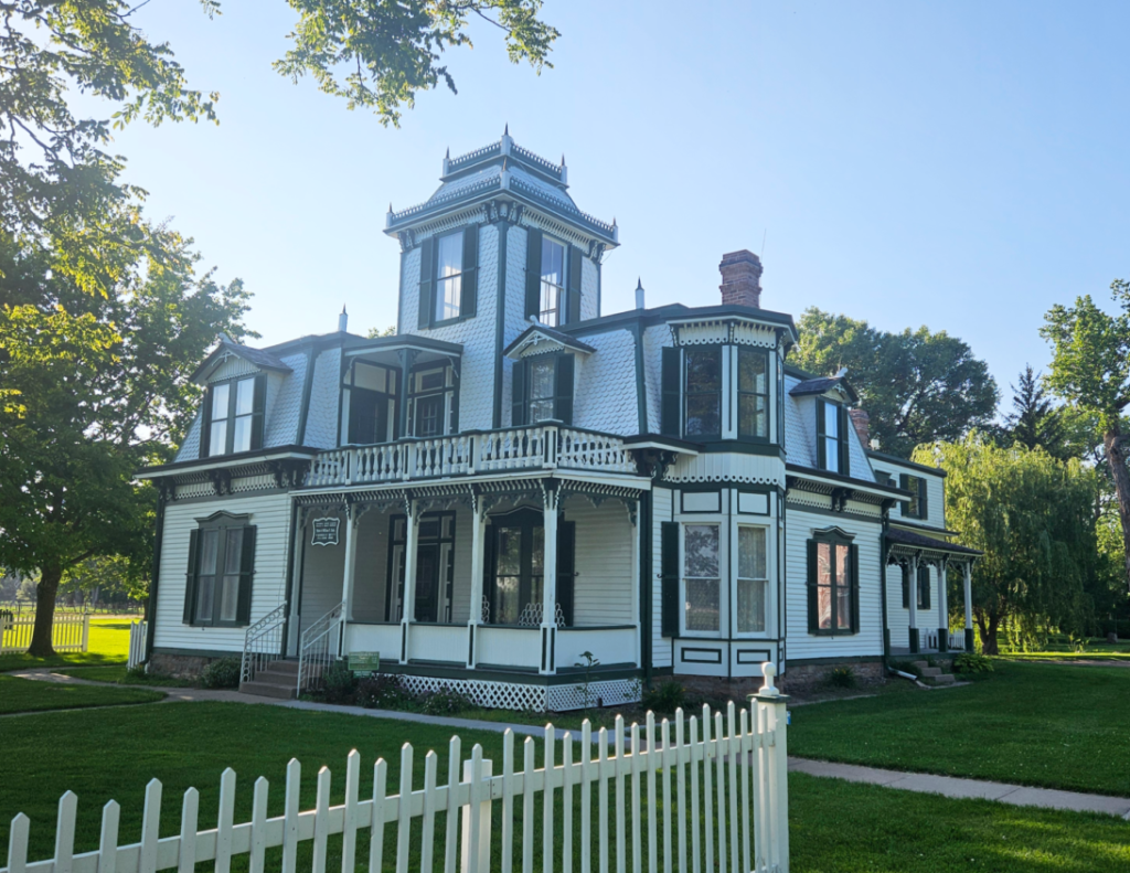 A two-story Victorian-style house with a white exterior, green trim, and a prominent tower, surrounded by a white picket fence and green lawn under a clear sky.