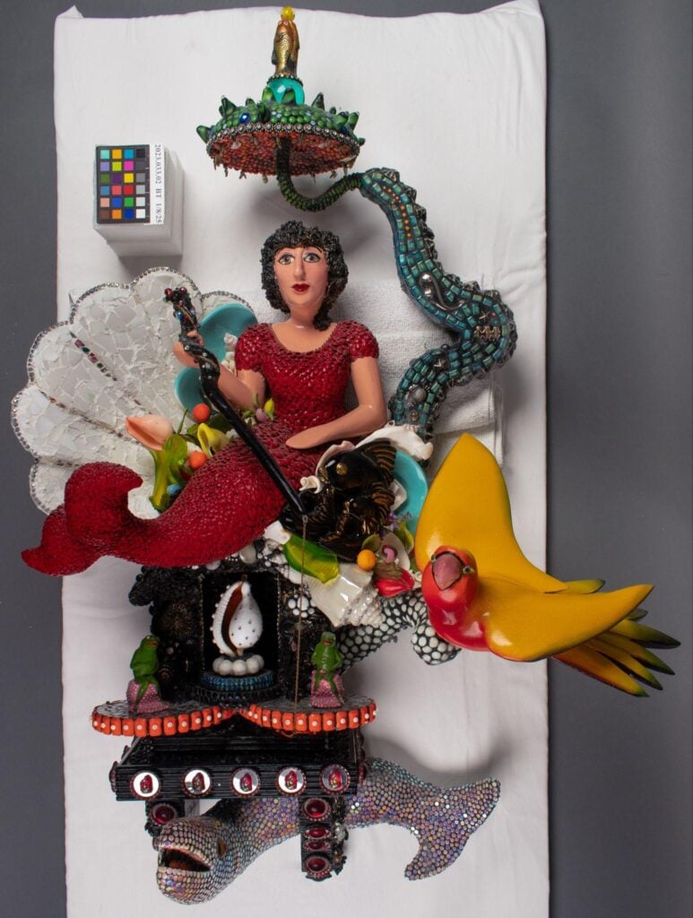 A mixed-media sculpture featuring a woman in red, a yellow bird, a clock, a shark, and various decorative elements, displayed on a white surface with a color calibration card.