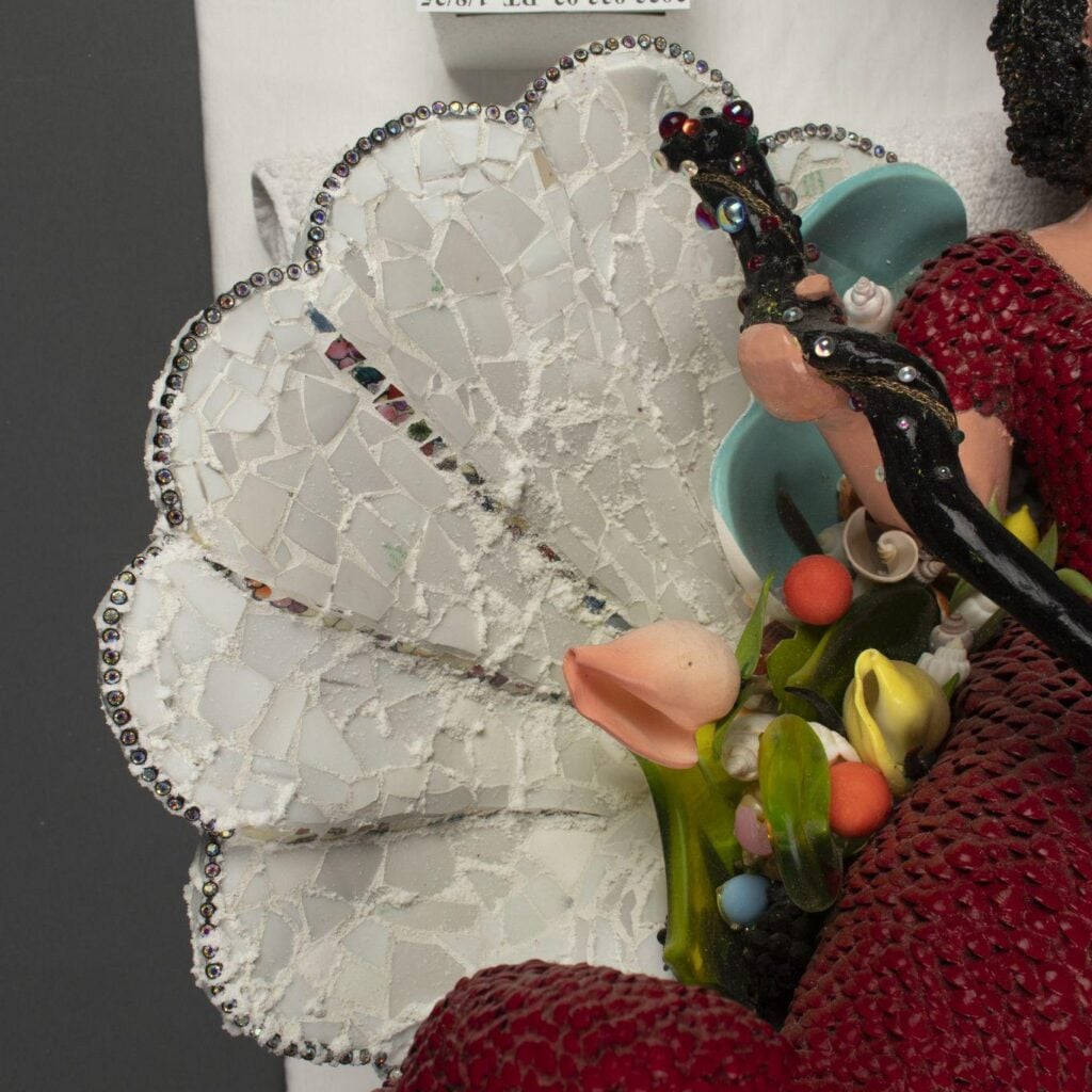 A close-up of a decorative mosaic featuring a fan made of white tiles with a beaded edge, artificial flowers, and a figure in a red textured garment.