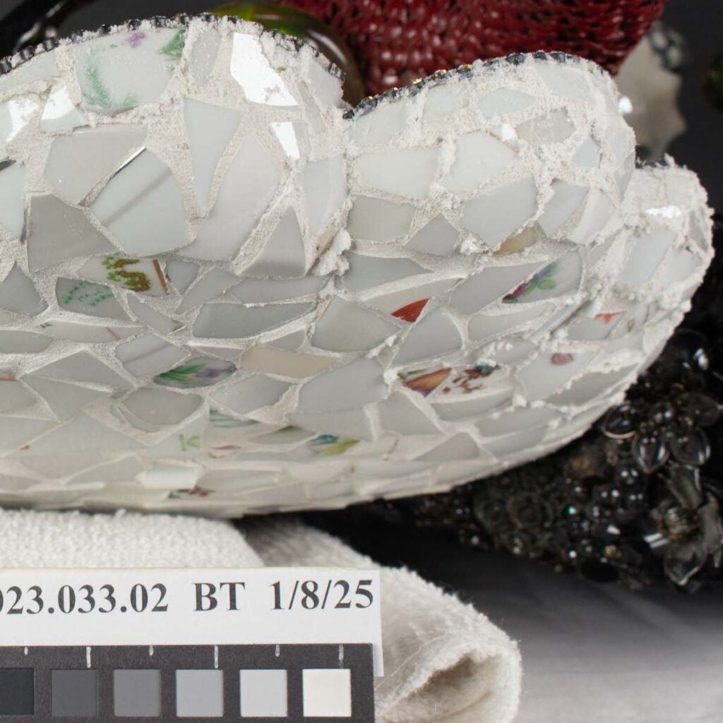 Close-up of a white, mosaic-patterned ceramic object with fragments of dishes, next to a rolled towel and a color calibration chart labeled "23.033.02 BT 1/8/25.