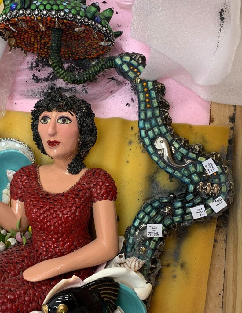 A mosaic sculpture of a woman in a red dress is lying on foam, with a green mosaic tentacle featuring small labeled tags extending above her.