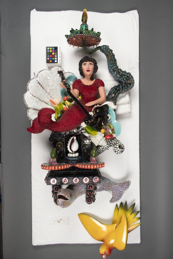 A colorful mixed-media sculpture featuring a woman in red, surrounded by various textured objects, animals, and a fish, displayed on a white cloth background.
