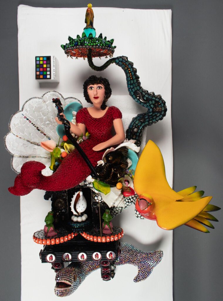 A colorful, detailed sculpture featuring a woman in red, birds, a fish, and various decorative elements is displayed on a white background. A color calibration card is visible in the corner.