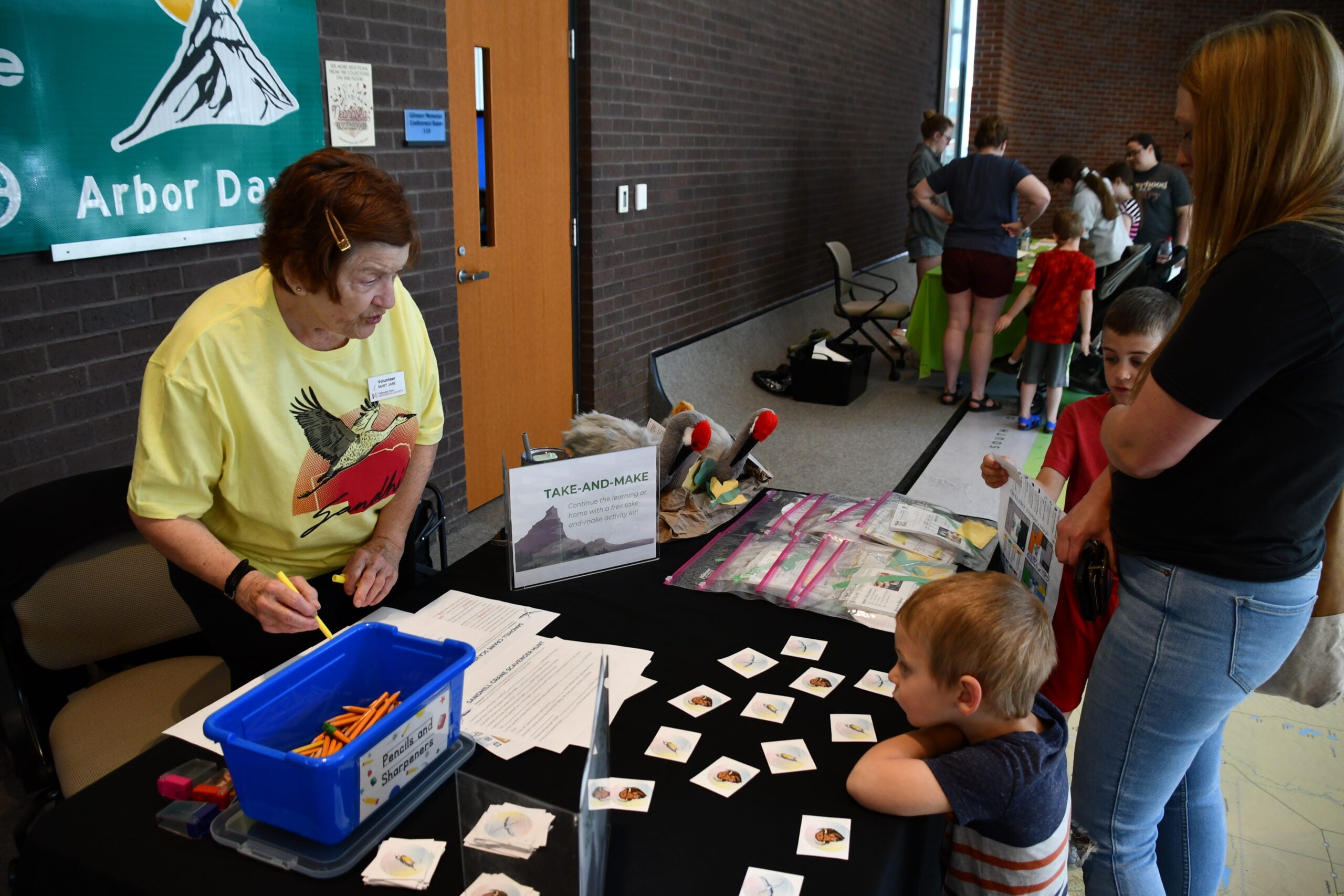An older woman at a table labeled "Take-and-Make" helps children and an adult with nature-themed activities at an indoor event. Other families are visible in the background.