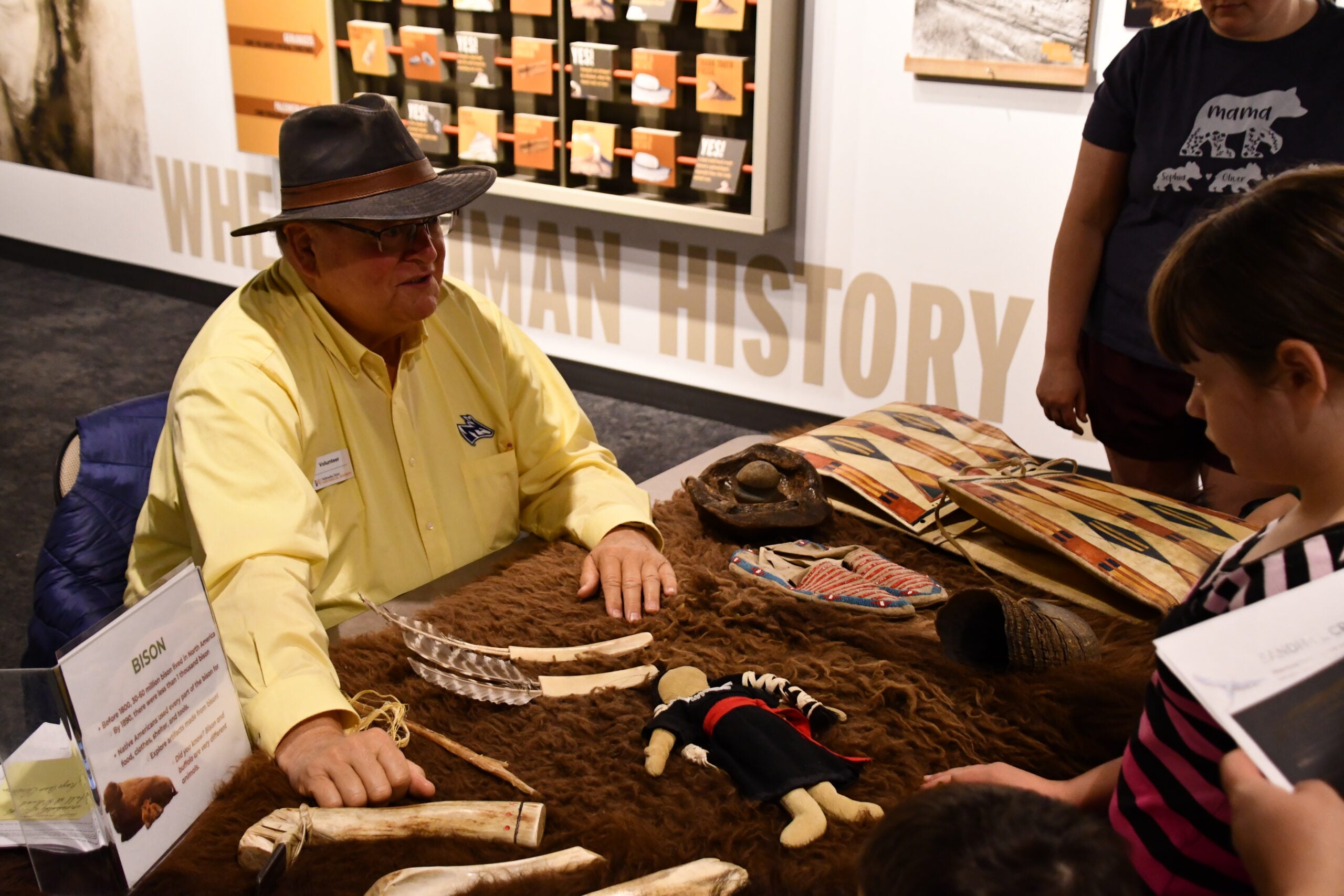 A man in a yellow shirt and hat sits at a table displaying Native American artifacts, talking to children at a museum exhibit.