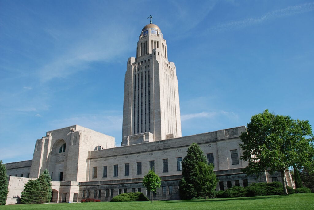 The Nebraska State Capitol building features a tall central tower with a golden dome, surrounded by landscaped lawns and trees under a clear blue sky.