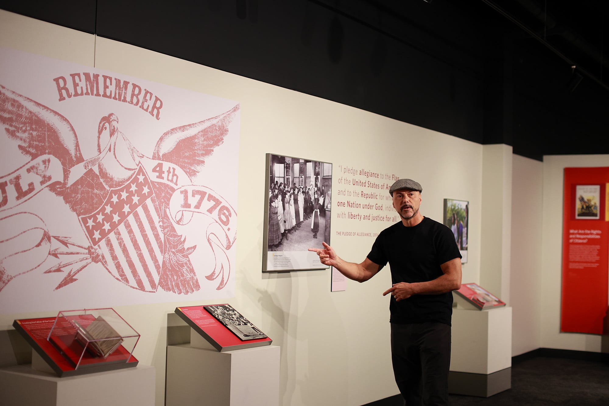 A man in a black shirt and cap stands in a museum, pointing to a historical display about July 4th, 1776, with informational panels and images on the wall.