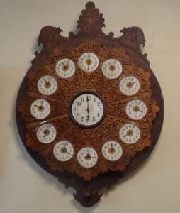 Ornate wooden wall clock featuring a large central clock face surrounded by twelve smaller clock faces, each numbered and set within decorative scrollwork.