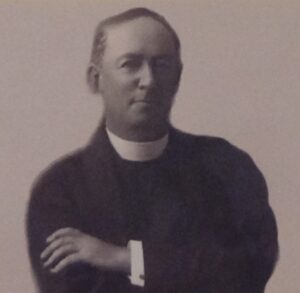 A man wearing clerical clothing with a high collar poses for a formal portrait, arms crossed, looking towards the camera.