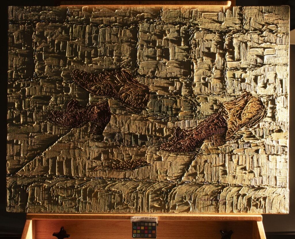 Textured abstract painting with gold and brown tones, featuring thick, layered brushstrokes and three indistinct shapes on a wooden easel.