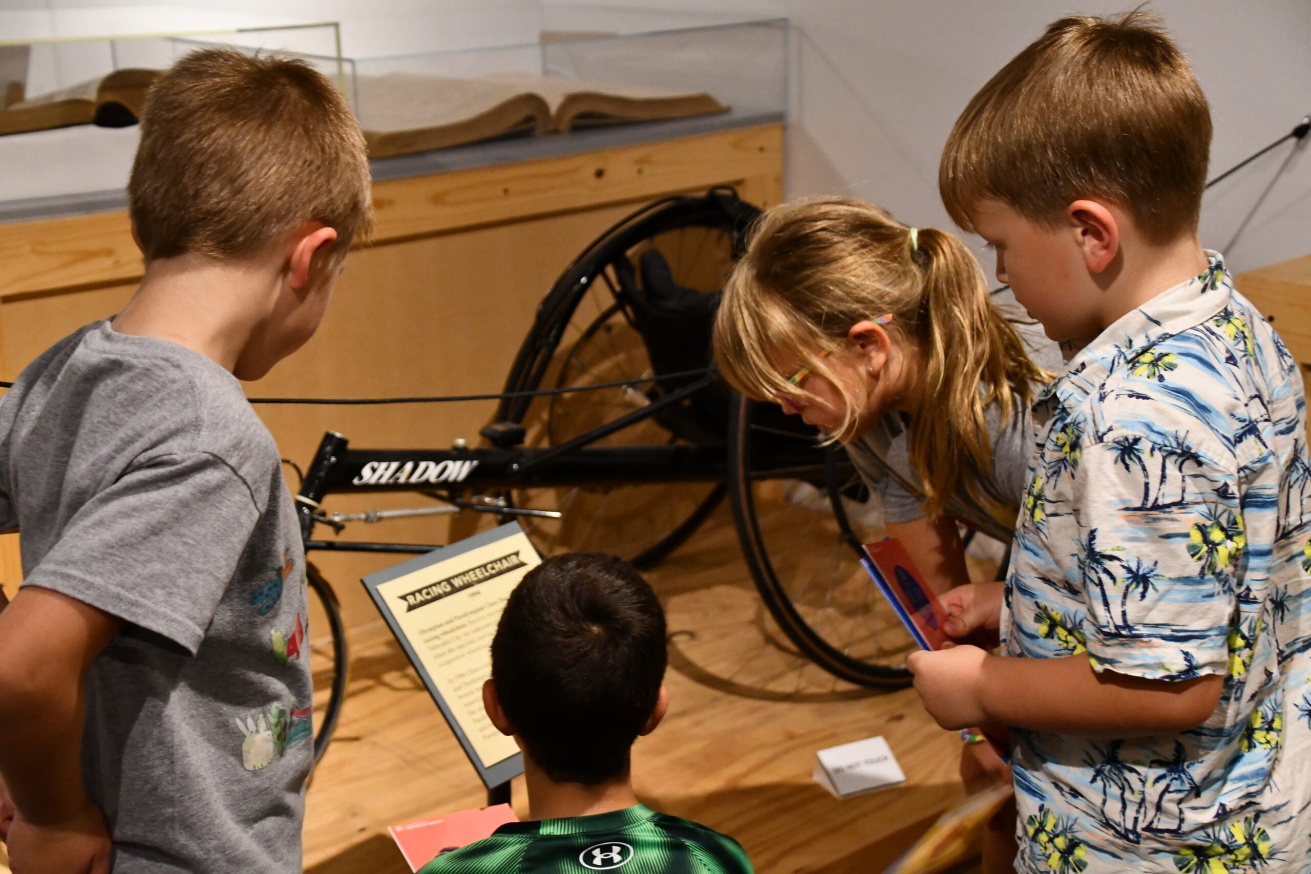Four children read an informational display about a wheelchair labeled "Shadow" in a museum exhibit during one of the museum's Summer Workshops.