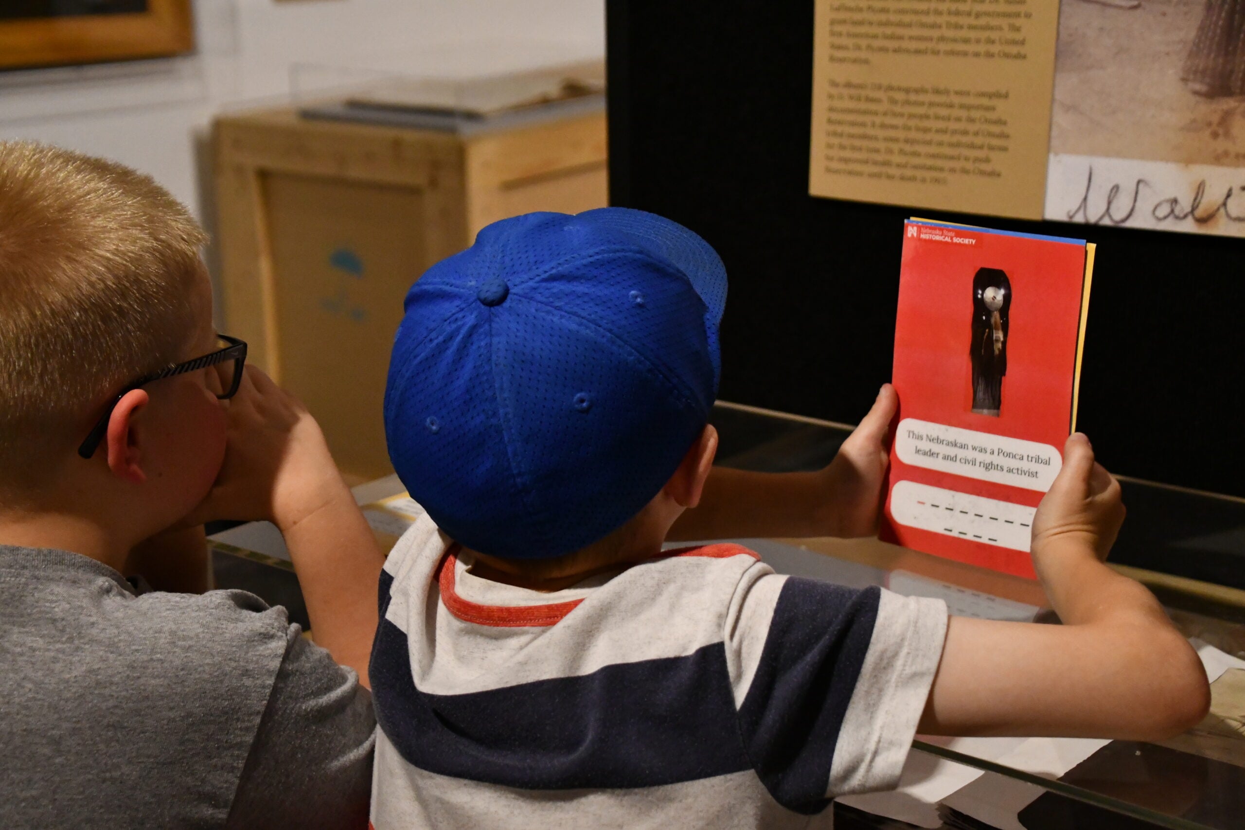 Two boys sit side by side at a display during the Summer Workshops, with one holding an informational card featuring an artifact and text.