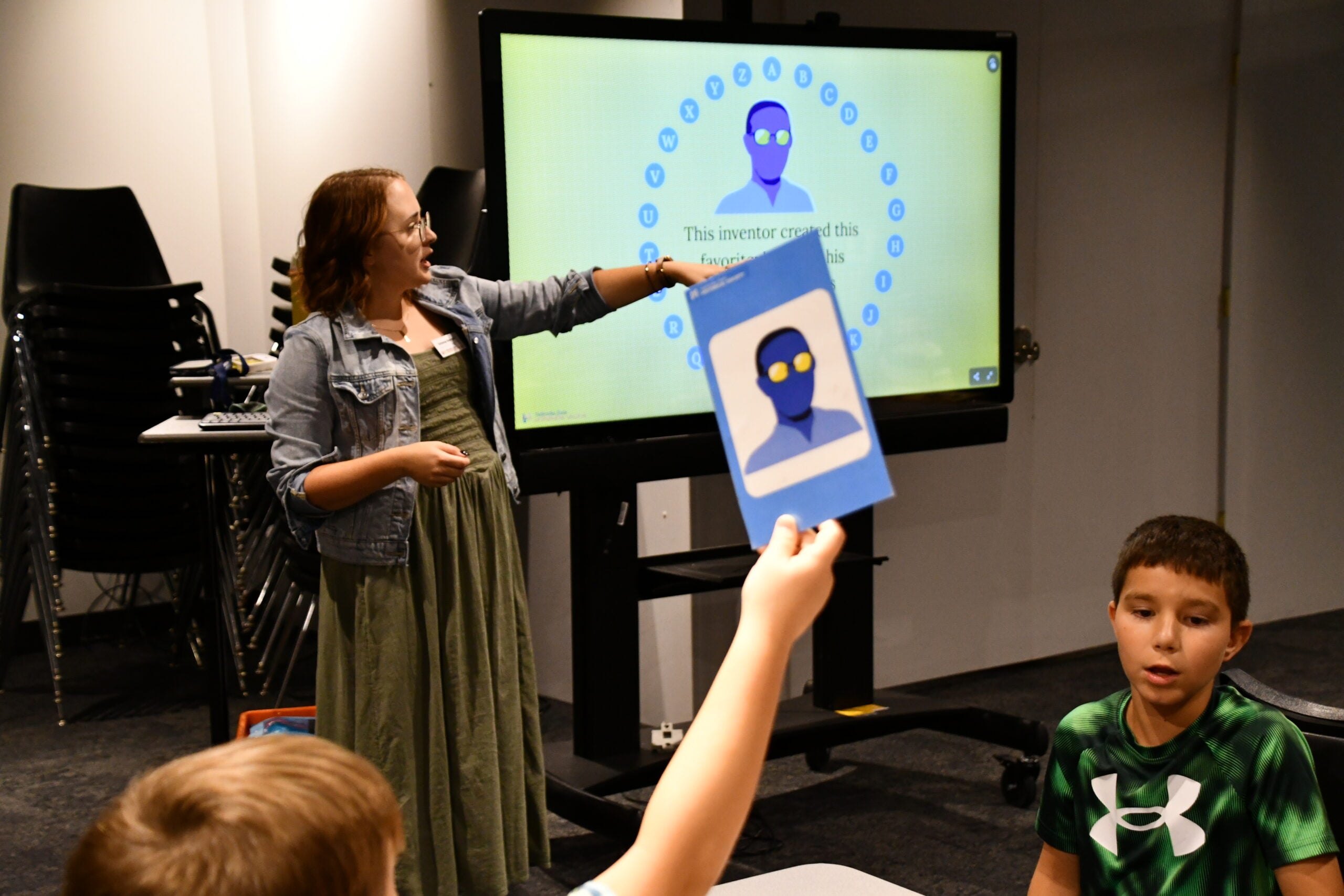 A woman stands near a digital screen with a quiz during Summer Workshops, pointing at it, while a student raises a card featuring a face illustration in a classroom setting.