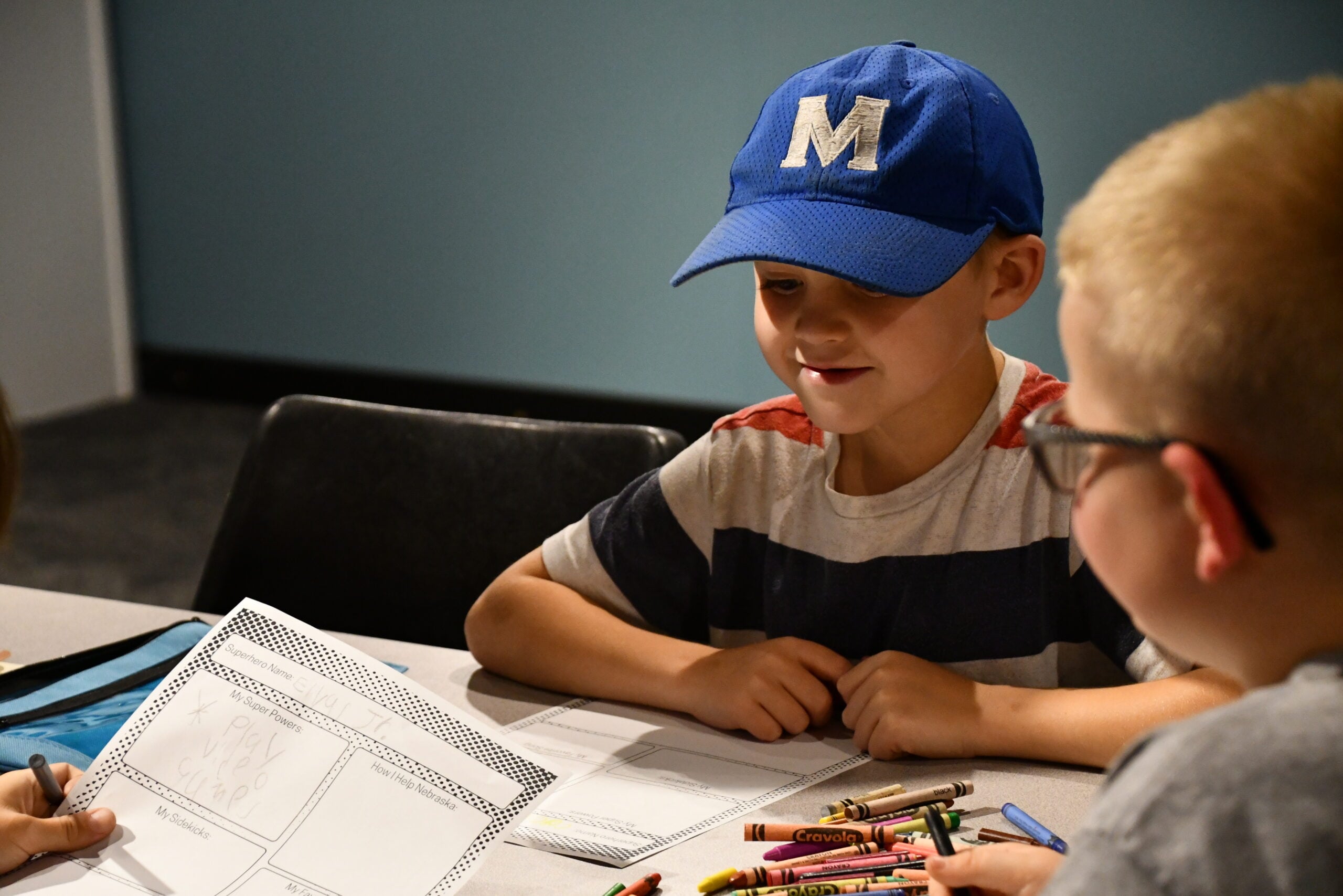 Two boys sit at a table during Summer Workshops, drawing with crayons on paper; one wears a blue "M" cap, the other wears glasses.