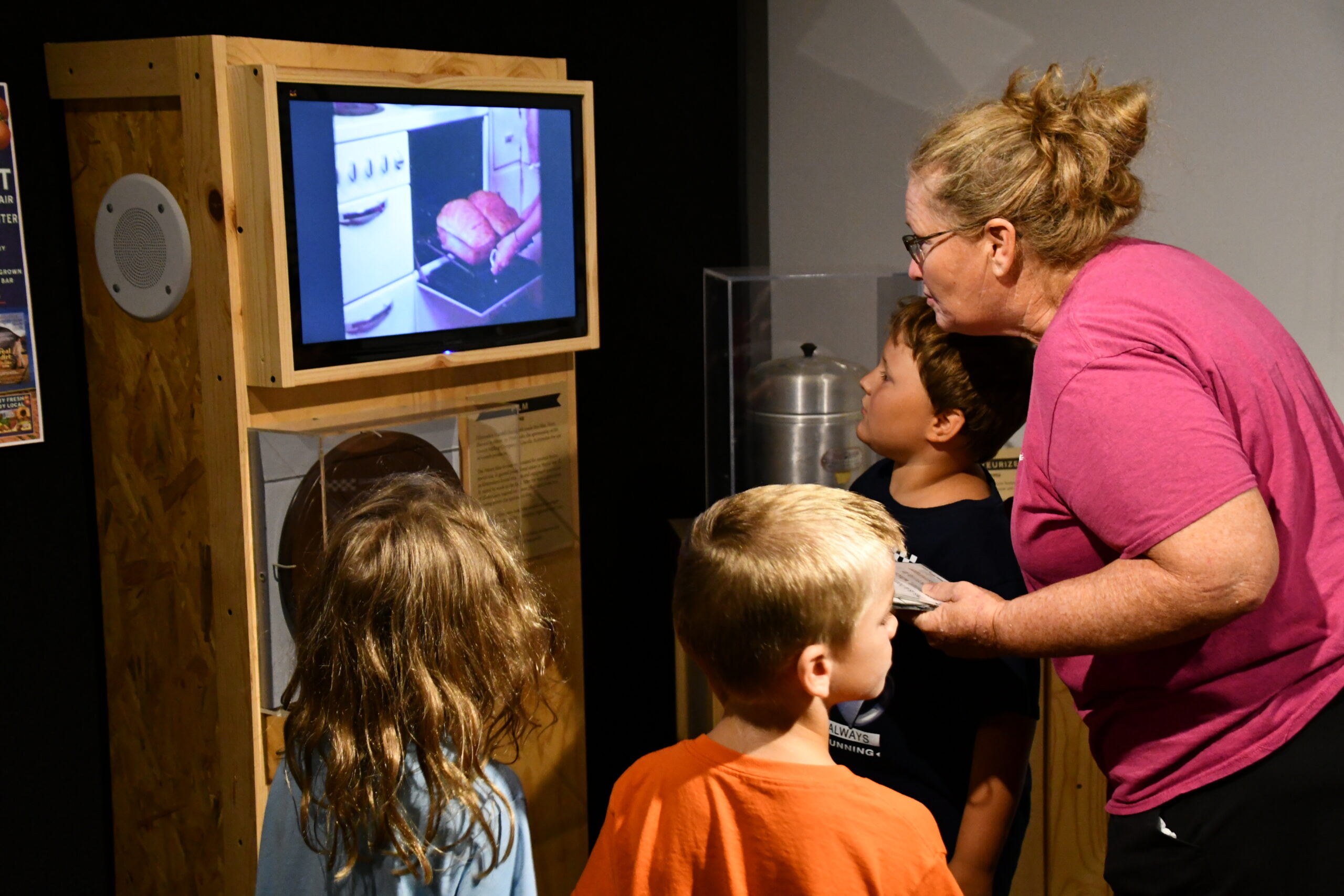 A woman and three children watch a video about food preparation on a screen at a museum exhibit, discovering ideas perfect for Summer Workshops.