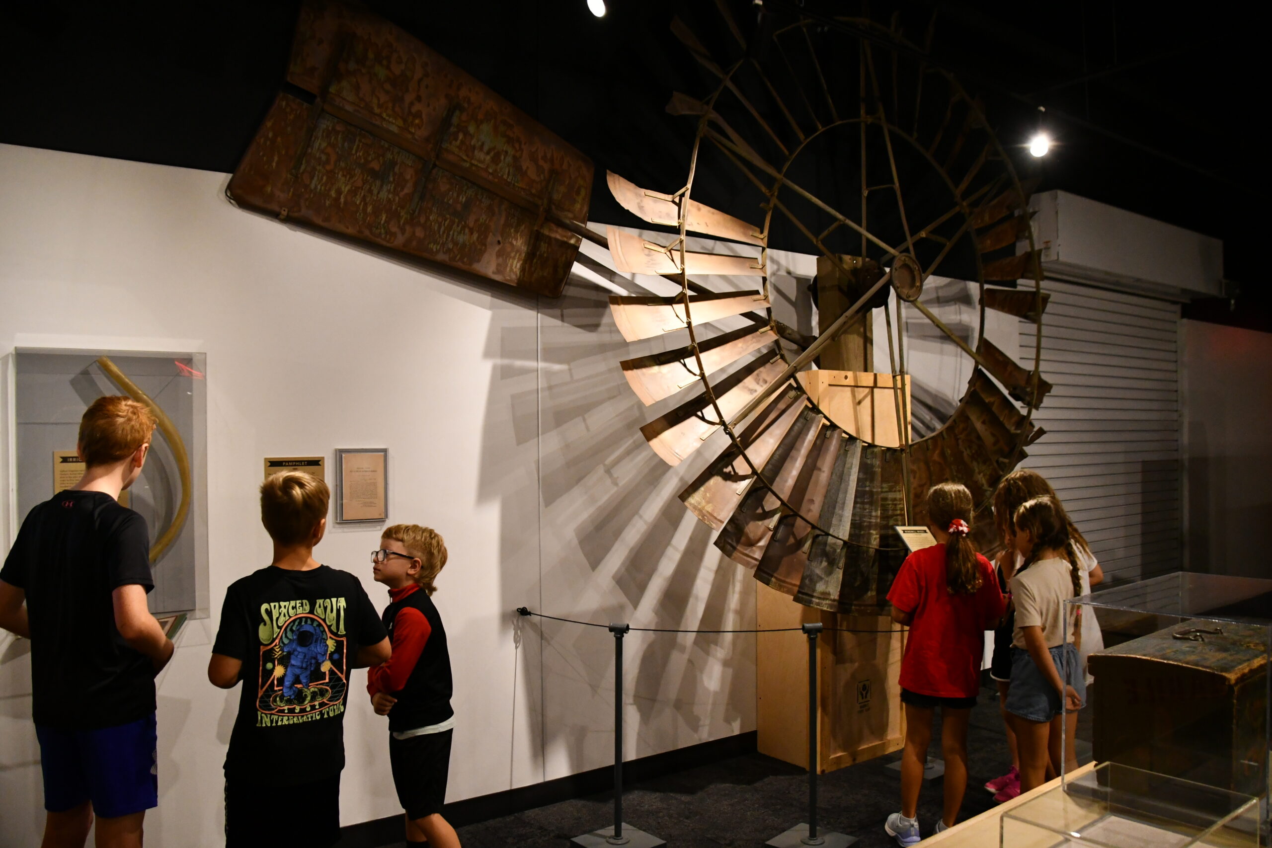 Children observe museum displays, including a large windmill structure mounted on the wall, in a well-lit exhibition room—part of the engaging Summer Workshops offered by the museum.