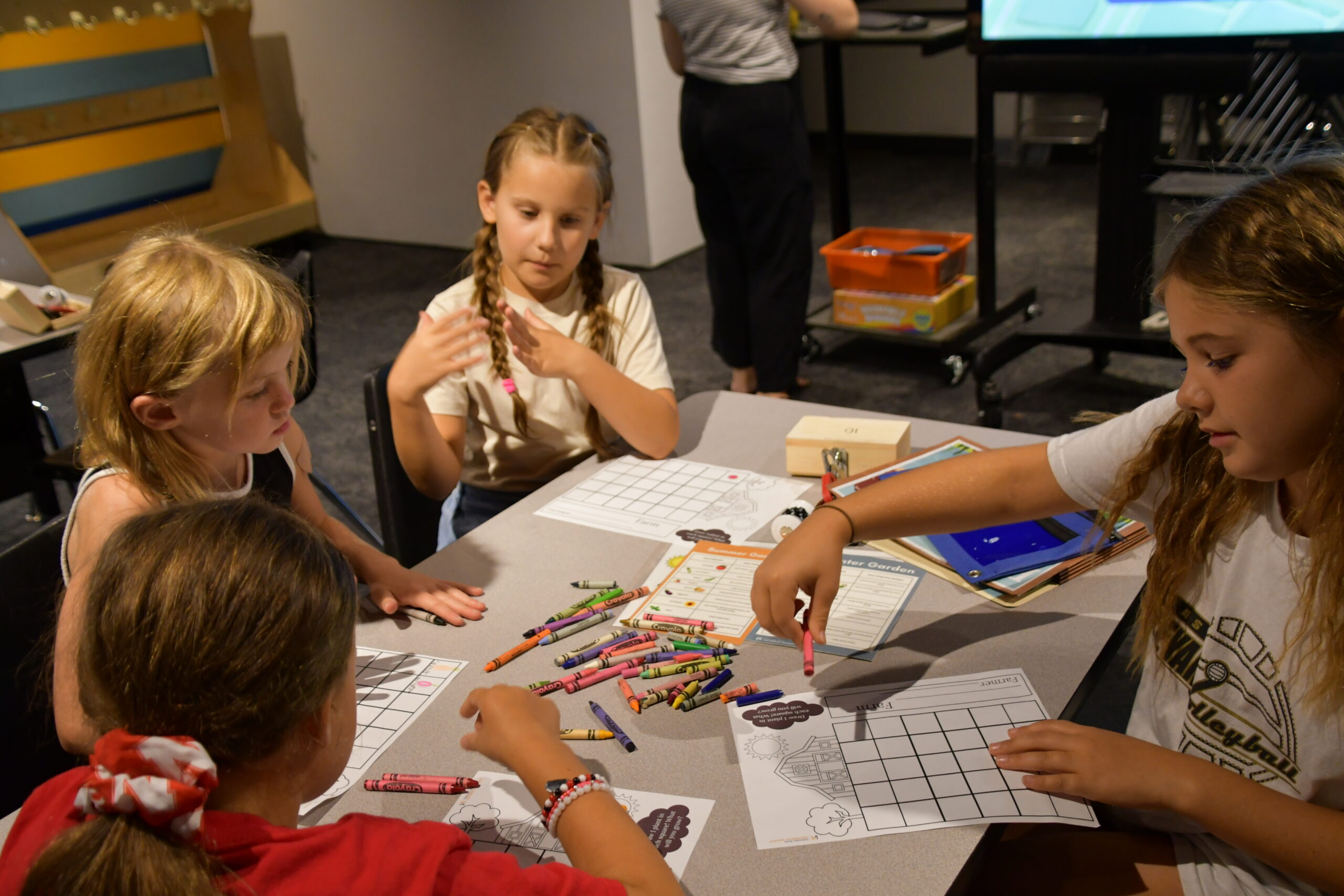 Four children sit around a table working on coloring sheets with crayons during Summer Workshops, while another person stands in the background.