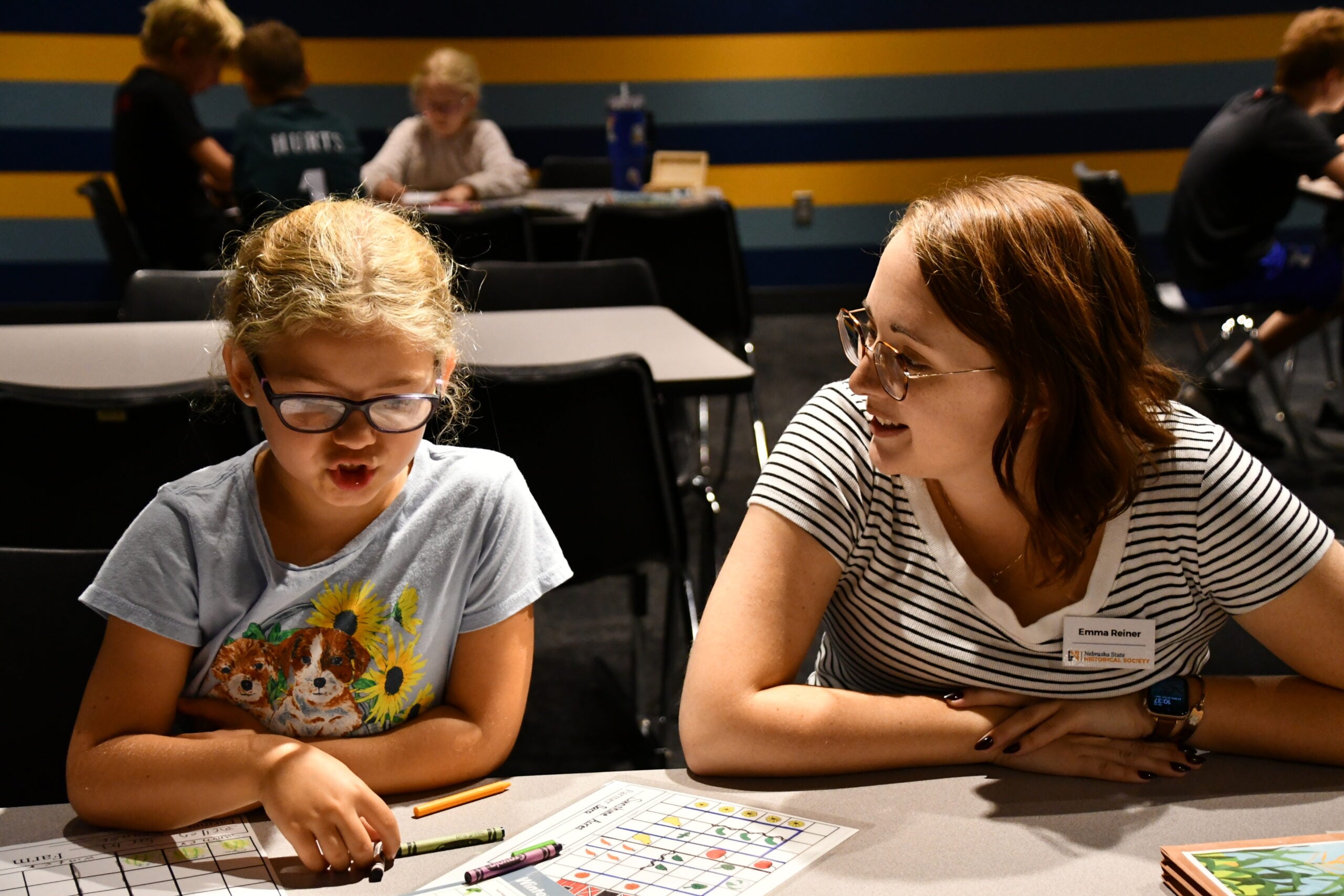 A young girl works on a worksheet with a woman wearing glasses and a name tag seated beside her, participating in Summer Workshops in a classroom with other students in the background.