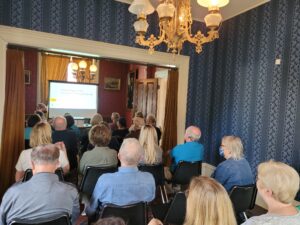 A group of people sit in a small room facing a screen displaying a presentation titled "Archaeology of the House of Seven Gables House.