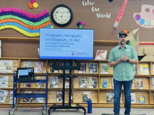 A man stands next to a display screen showing a presentation titled “Pictographs, Petroglyphs, and Geoglyphs, Oh My!” in a room decorated with colorful art and bookshelves.